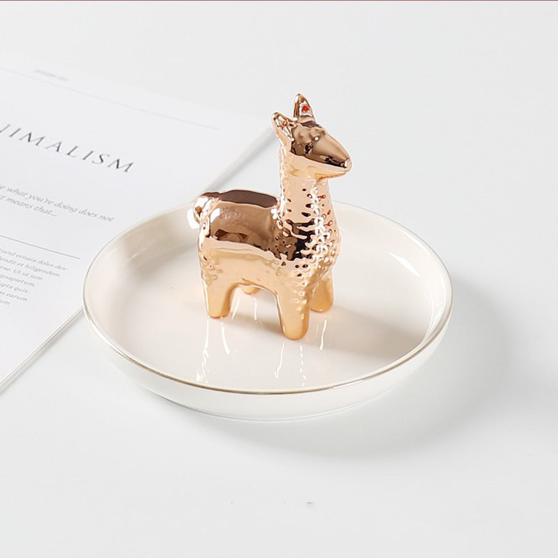 White ceramic jewelry tray with gold rim, and a rose golden alpaca standing on the middle of the dish. 