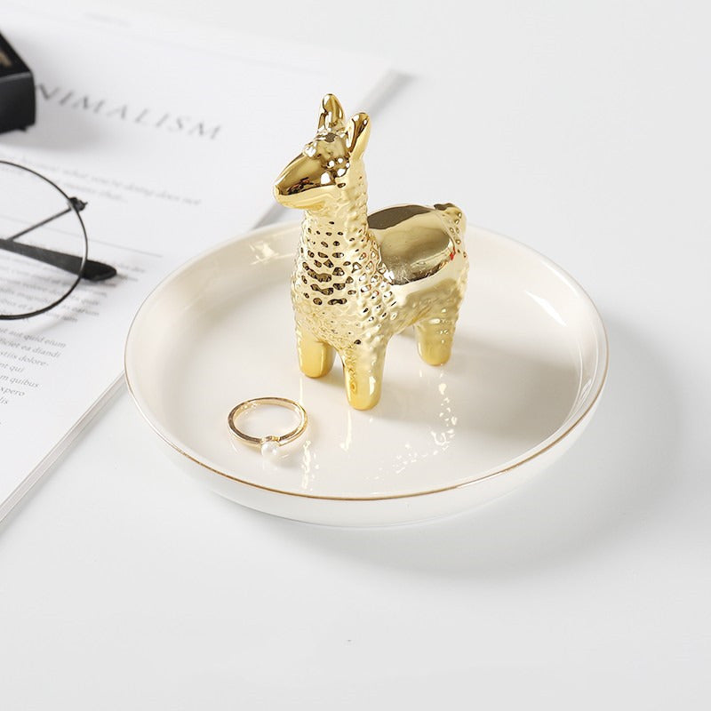 White ceramic jewelry tray with gold rim, and a golden alpaca standing on the middle of the dish.