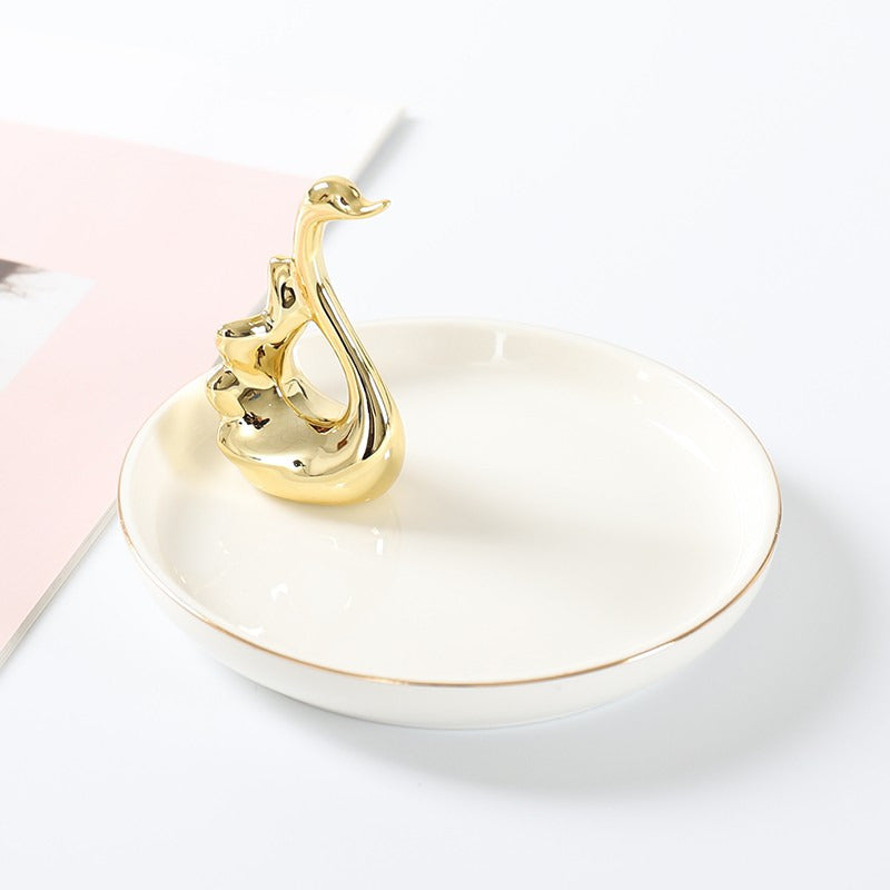 White ceramic jewelry tray with gold rim, and a golden swan sitting on the edge of the dish.