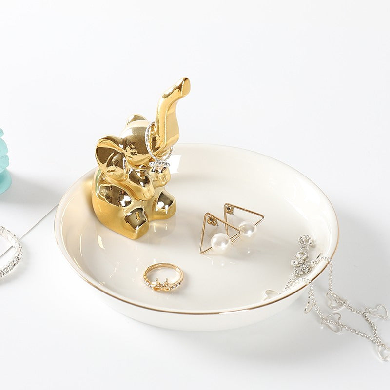 White ceramic jewelry tray with gold rim, and a golden elephant sitting on the edge of the dish.