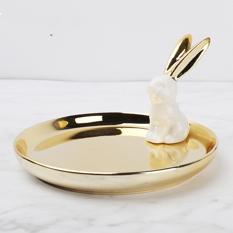 Golden ceramic jewelry tray, and a white rabbit  with golden ears sitting on the edge of the dish.